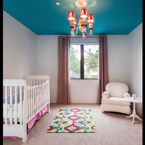 以下が含まれることがあります： 青い天井とカラフルな敷物のある子供部屋に、ピンクのフリルが付いた白いベビーベッドと、サイドテーブルのある白いアームチェアがある。