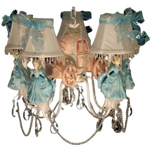 May include: A white chandelier with crystal accents features a pink Cinderella carriage and four blue Cinderella figurines. Each light fixture has a white lampshade with a blue ribbon.