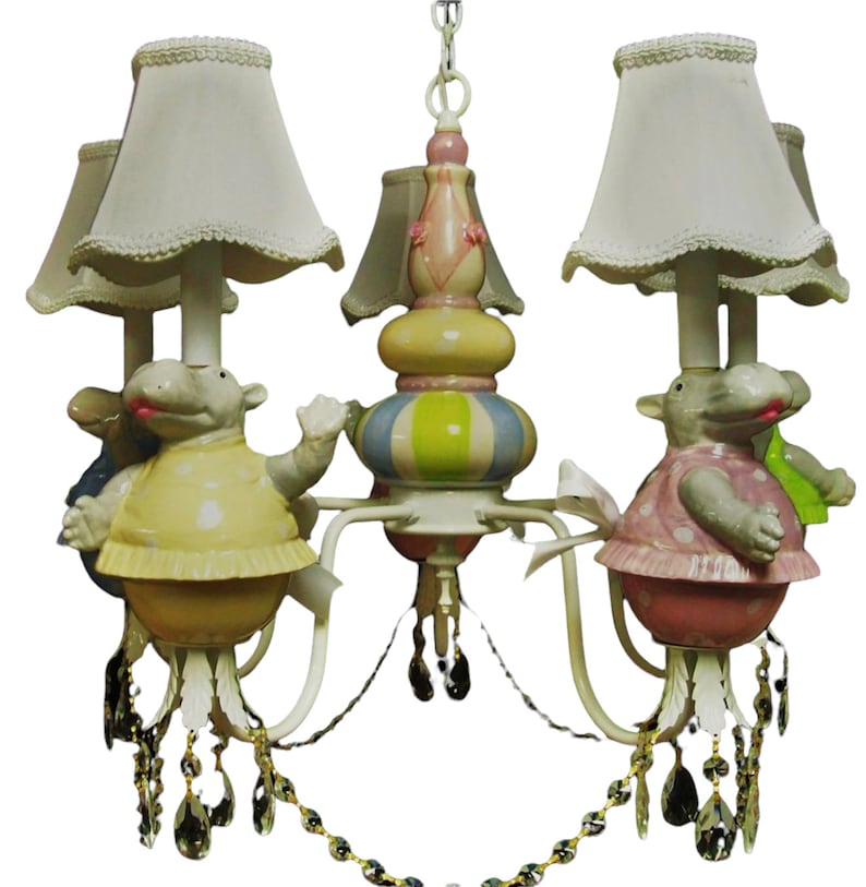 May include: A white chandelier with four light bulbs and white lampshades. The chandelier features four ceramic hippopotamus figurines with polka dot dresses, each holding a light bulb. The chandelier has crystal accents.