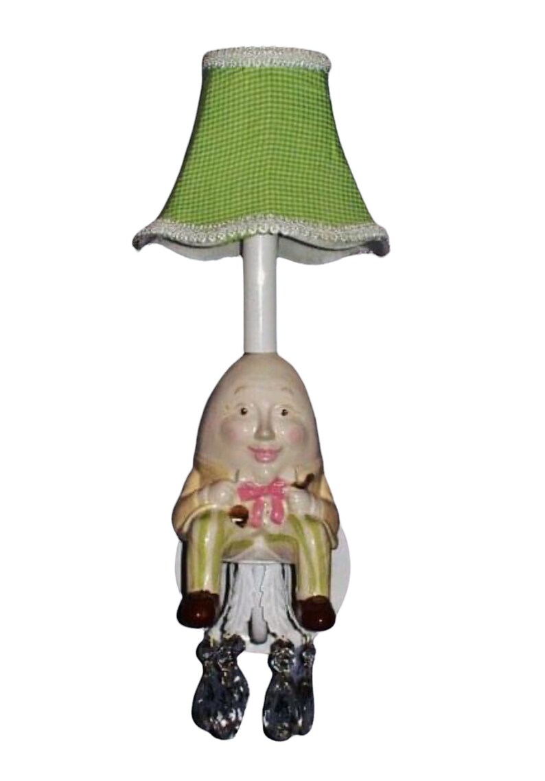 May include: A green gingham lampshade with a white trim sits atop a ceramic lamp base shaped like a seated Humpty Dumpty figure. The figure has a white face with painted features, a pink bow tie, and green and white striped pants. The lamp base has crystal-like accents on the feet.