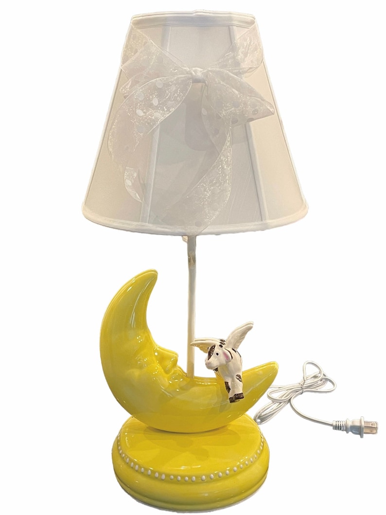 May include: A yellow ceramic table lamp shaped like a crescent moon with a white pig with wings sitting on top. The lamp has a white fabric shade with a white bow.