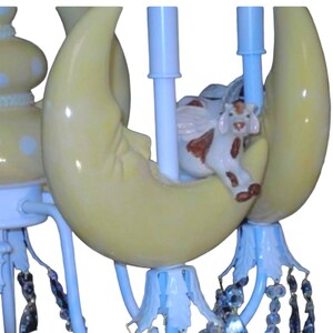K&ouml;nnte beinhalten: Ein wei&szlig;er und gelber Keramik-Kronleuchter mit einem Halbmond-Design und einer kleinen Kuhfigur. Der Kronleuchter hat Kristall-Akzente und ist perfekt f&uuml;r ein Kinderzimmer oder ein Kinderzimmer.