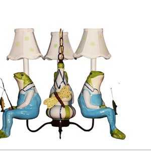 May include: A three-light chandelier with frog figurines holding fishing poles and a dragonfly design on the center light. Each frog is wearing blue overalls and has a green and white patterned body. The lampshades are white with green polka dots.