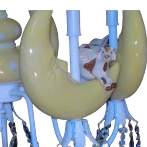 May include: A white and yellow ceramic chandelier with a crescent moon design and a small cow figurine sitting on top. The chandelier has crystal-like beads hanging from the bottom.