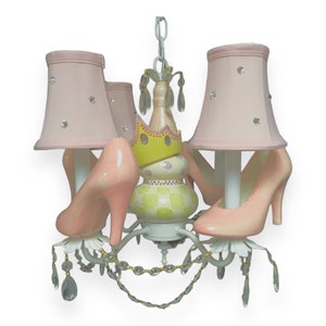 May include: A white chandelier with pink lampshades and pink high heel shoes. The chandelier has a yellow and green crown in the center and crystal accents.