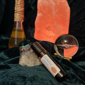 May include: A brown glass roller bottle with a white label that says "Grandmas House" and a picture of a pie. The bottle is sitting on a clear crystal with a pink Himalayan salt lamp in the background.