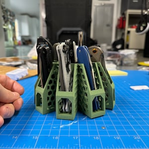 Puede incluir: Un organizador de cuchillos de plástico verde de múltiples ranuras con un patrón de panal. El organizador contiene una variedad de cuchillos de bolsillo con mangos de diferentes colores.