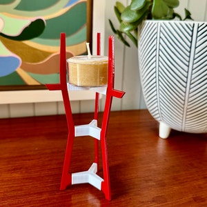 May include: A red metal candle holder with a white base and a single tea light candle. The candle holder is shaped like a tower with a triangular base.