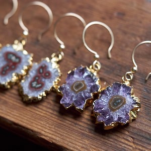 Puede incluir: Cuatro pares de pendientes de oro con detalles de piedras de amatista y ágata. Los pendientes tienen forma de flor con un bucle de alambre dorado.