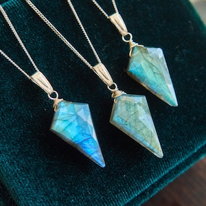 May include: Three gemstone pendants with silver chains rest on a dark green velvet surface. Each pendant features a faceted, kite-shaped labradorite stone, displaying shades of blue and green. The necklaces are displayed close together, showcasing the unique color variations.