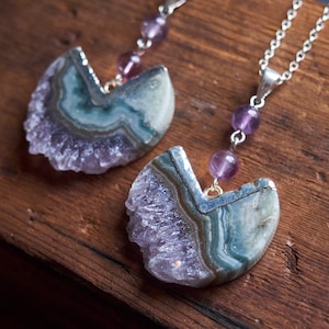 May include: Two silver necklaces with amethyst crystal pendants. The pendants are half-circle shaped and have a layered, agate-like pattern in shades of purple, green, and white. The necklaces are adorned with small, purple amethyst beads.
