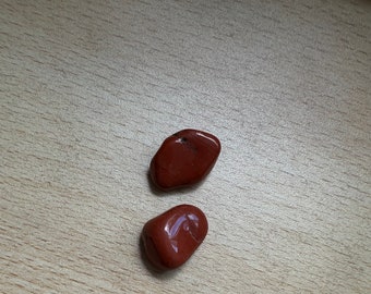 May include: Two smooth, red stones on a light wood surface. The stones are irregularly shaped and have a glossy finish.