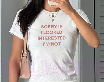 May include: White t-shirt with the text "SORRY IF I LOOKED INTERESTED I'M NOT" in red. The shirt is short-sleeved and has a crew neck. The person is wearing sunglasses and a necklace.