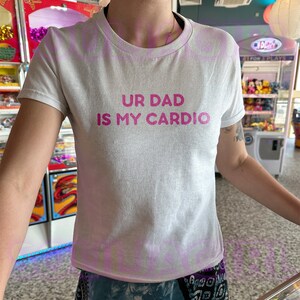 May include: White t-shirt with pink text that reads "UR DAD IS MY CARDIO"