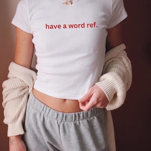 Peut inclure: T-shirt blanc à manches courtes avec le texte rouge "have a word ref.". Le haut est associé à un pantalon de survêtement gris et un cardigan crème. Un collier doré est visible. L'ensemble est décontracté et confortable.