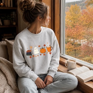 Peut inclure: Un sweat-shirt gris clair à col rond orné d'un motif imprimé représentant une pile de livres, un petit fantôme blanc, une citrouille orange et une tasse de boisson chaude, le tout entouré de feuilles d'érable automnales orange et jaunes.