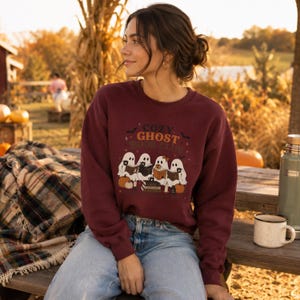 Peut inclure: Un sweat-shirt à col rond et manches longues bordeaux avec une impression graphique représentant quatre fantômes blancs lisant des livres. Le texte "COZY GHOST BOOK CLUB" est imprimé au-dessus, avec des citrouilles, des étoiles et des chauves-souris.