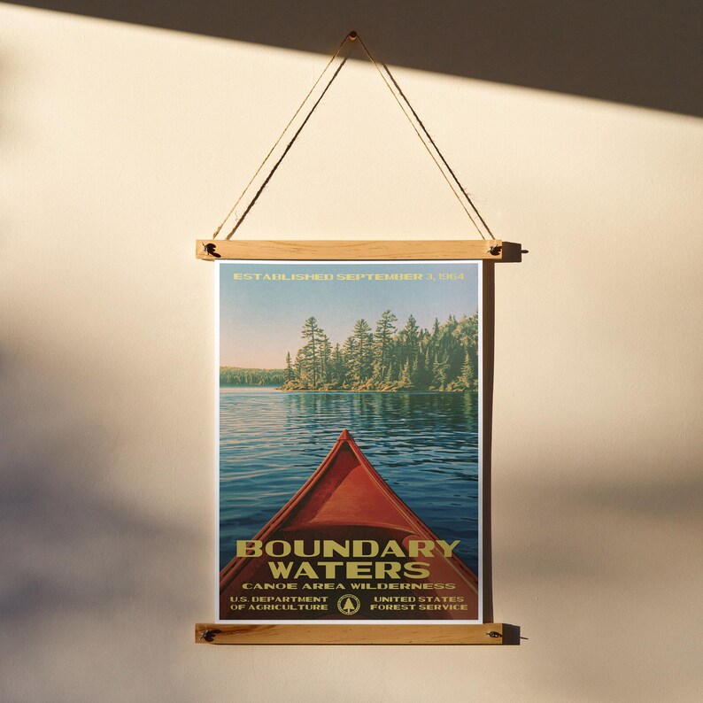 Peut inclure: Une affiche verticale montrant la proue d'un cano&euml; rouge sur une eau bleue, avec une for&ecirc;t en arri&egrave;re-plan et le texte "BOUNDARY WATERS CANOE AREA WILDERNESS" et "ESTABLISHED SEPTEMBER 3, 1964". L'affiche est fix&eacute;e par deux baguettes en bois clair.
