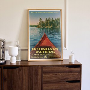Peut inclure: Une affiche encadr&eacute;e montrant un cano&euml; rouge sur une eau bleue avec une for&ecirc;t en arri&egrave;re-plan. Le texte indique : ESTABLISHED SEPTEMBER 3, 1964, BOUNDARY WATERS CANOE AREA WILDERNESS, U.S. DEPARTMENT OF AGRICULTURE, UNITED STATES FOREST SERVICE.
