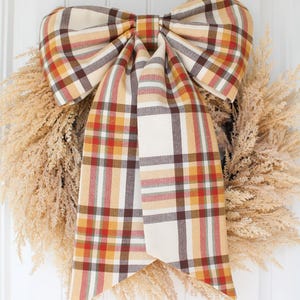 May include: A decorative wreath made of light tan, feathery dried foliage, featuring a large, prominent fabric bow tied at the top. The bow is patterned with a plaid design in shades of cream, mustard yellow, dark brown, and muted red.