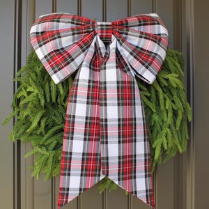 May include: A large decorative bow made of fabric with a red, white, black, and gray plaid pattern. The bow features two wide loops and two long, hanging tails, and it is positioned over a circular wreath made of green pine needles.