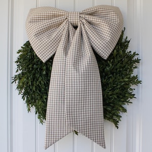 May include: A green wreath with a large, tan and white gingham bow. The bow is centered on the wreath and has long, flowing tails. The wreath is hanging on a white door.