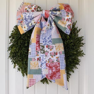 May include: A decorative wreath with a large, patchwork fabric bow. The bow features a variety of floral and patterned fabrics in red, blue, yellow, and green. The wreath is made of green foliage and hangs on a white door.