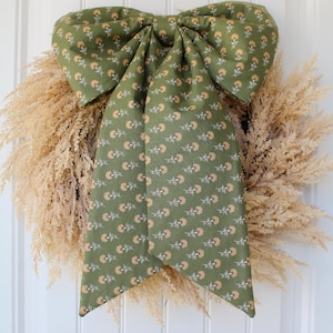 May include: A decorative wreath made of dried, light beige pampas grass featuring a large, fabric bow centered on top. The bow is olive green with a repeating pattern of small, light orange flowers and white stems, featuring two long tails.