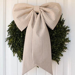 May include: A large decorative bow made of beige and white gingham fabric featuring two wide loops and two long, straight-edged tails. The bow is positioned over a circular wreath composed of dense, dark green leaves with small, rounded foliage.