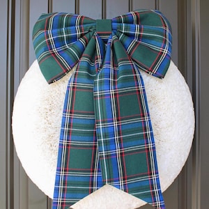 May include: A large decorative bow made of fabric featuring a green, blue, and red plaid tartan pattern. The bow has two wide loops at the top and two long, hanging tails with angled ends, centered over a circular, textured white wreath base.