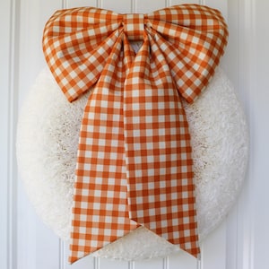 May include: A circular white wreath with a textured, fluffy surface adorned with a large, fabric bow featuring an orange and white gingham check pattern. The bow has two wide, long tails hanging down over the center of the round white wreath.