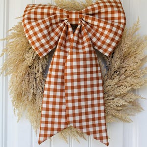 May include: A decorative wreath featuring a large, central bow made of fabric with a burnt orange and white gingham check pattern. The bow is layered over a circular base composed of dried, light beige, feathery botanical elements.