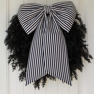 May include: A circular wreath made of dark black evergreen foliage, adorned with a large, prominent bow featuring a bold black and white vertical striped pattern. The bow has two wide loops and two long, hanging tails that drape over the center.