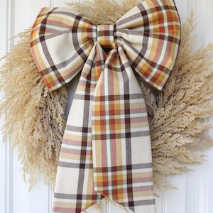 May include: A large decorative bow made of fabric featuring a plaid pattern in shades of cream, mustard yellow, burnt orange, and dark brown. The bow has two long, hanging tails and is positioned over a circular wreath made of dried beige grasses.