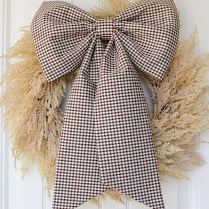 May include: A large decorative bow made of brown and white gingham fabric featuring two long, hanging tails, centered over a circular wreath constructed from dried, light beige feathery grasses, mounted on a white paneled door.
