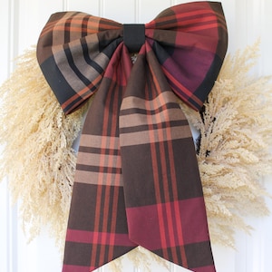 May include: A large decorative bow made of fabric featuring a plaid pattern in shades of dark brown, deep red, and muted orange. The bow has two long, wide tails hanging downward and is secured in the center with a solid black fabric band.