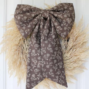 May include: A large, decorative bow made of brown fabric with a white floral pattern, hanging over a wreath of dried, beige grasses. The bow is centered on a white door.