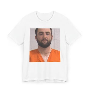 Puede incluir: Una camiseta blanca con una foto de un hombre con un mono naranja. El hombre tiene barba y pelo corto castaño.
