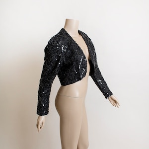 May include: A black sequined cropped jacket with long sleeves. The jacket is open in the front and has a V-neck.