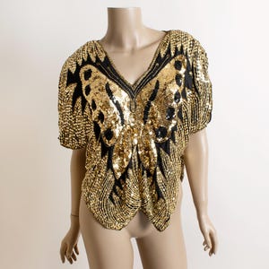 May include: A gold sequined top with a butterfly design. The top features a V-neck and short sleeves. The butterfly design is created with black and gold sequins, creating a striking contrast. The top is displayed on a mannequin.