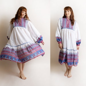 May include: A white maxi dress with a blue and red geometric pattern around the hem. The dress has long sleeves with a gathered cuff and a round neckline.