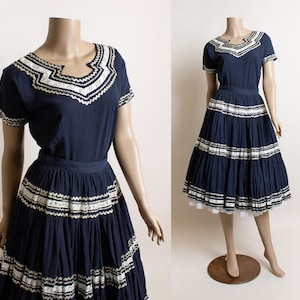May include: A navy blue dress with white and cream-colored embroidered trim. The dress features a short-sleeved top and a tiered skirt. The dress is displayed on a mannequin. The dress is a vintage style.