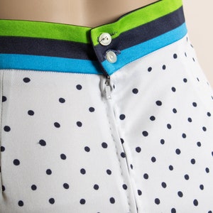 May include: A white skirt with black polka dots and a blue, green, and white striped waistband. The skirt has a zipper closure and two buttons at the top.