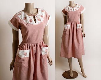 May include: A vintage-style, coral-colored dress with white accents. The dress features a unique collar and pocket design, with white trim and circular cutouts. The dress is displayed on a mannequin, showcasing its classic silhouette and details.