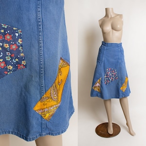 May include: A blue denim skirt with a patchwork design. The skirt has three patches, one with a floral pattern, one with a paisley pattern, and one with a yellow and orange pattern.