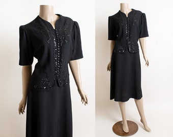 May include: A black two-piece suit with a beaded design on the jacket. The skirt is knee-length and the jacket has short sleeves.