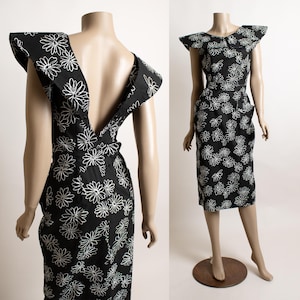 May include: A black midi dress featuring a white daisy floral print, a wide collar with extended shoulder flaps, a deep V-neckline, a fitted waist, and a straight skirt silhouette. The dress is displayed on a mannequin showing both front and back views.