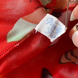 May include: A white fabric tag with blue lettering that reads "Jolie" attached to a red and white floral fabric.