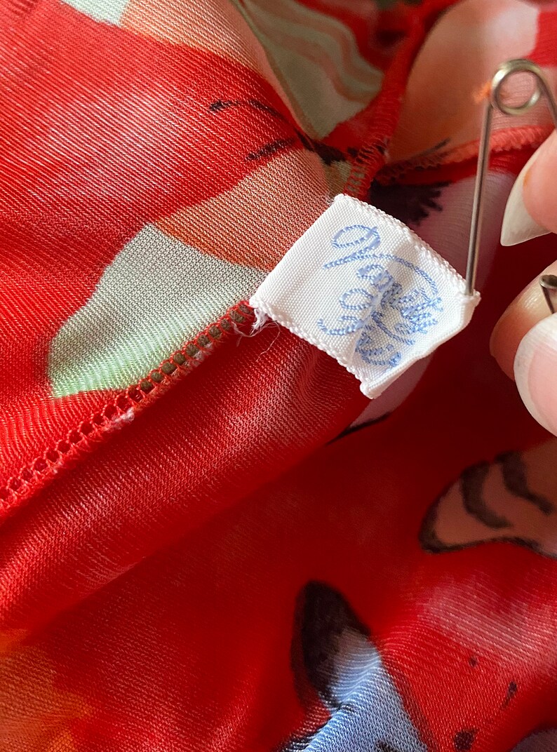 May include: A white fabric tag with blue lettering that reads "Jolie" attached to a red and white floral fabric.