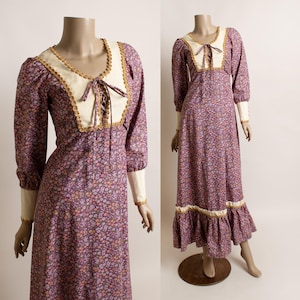 May include: A long-sleeved floor-length dress featuring a purple floral print, a cream-colored bodice panel with a lace-up front, and gold trim. The garment includes cream cuffs, a ruffled hemline, and a fitted waist, displayed on a mannequin.
